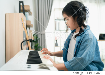 Young girl plays piano at home in cozy living room while wearing a denim jacket and focusing on her musical practice and creativity development 139325876