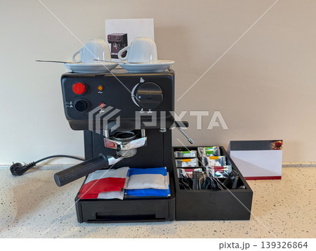 Coffee machine with cups and condiments on countertop in hotel room interior with power outlet, wall background. Hospitality service, travel accommodation, business stay, guest comfort, hotel amenity 139326864