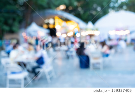 Blurred image of crowded outdoor market with people sitting at tables and food stalls in background in park 139327409