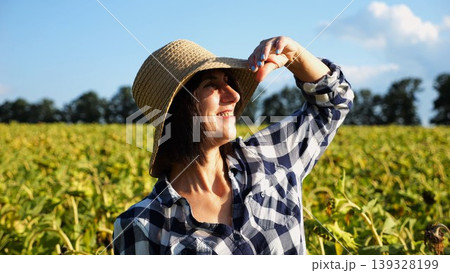 Happy smiling female farmer looks into sunflower field and enjoys harvest. Portrait of adult beautiful agronomist in straw hat with yellow plantation at background. Concept of agricultural business Happy smiling female farmer looks into sunflower field and enjoys harvest. Portrait of adult beautiful agronomist in straw hat with yellow plantation at background. Concept of agricultural business 139328199