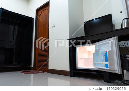 Low angle view of the opened mini fridge with water bottles placed under wooden table near the curtains in a hotel room. 139329066