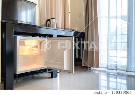 Low angle view of the opened mini fridge with water bottles placed under wooden table near the curtains in a hotel room. Low angle view of the opened mini fridge with water bottles placed under wooden table near the curtains in a hotel room. 139329069