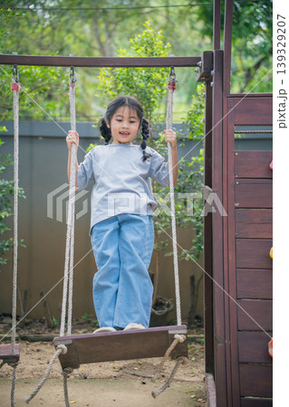 Young girl on swing enjoying outdoor playtime in nature, wearing casual clothing, smiling joyfully in a playground setting surrounded by greenery 139329207