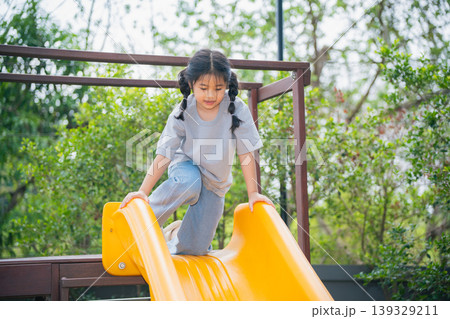 Happy child playing on yellow slide in outdoor playground, enjoying sunny day with trees and greenery in background, carefree childhood fun and laughter 139329211