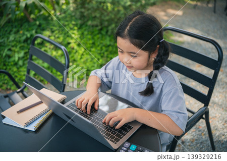 Young girl engaged in focused work on laptop outdoors, sitting at table with notepad and pencil in serene garden setting with lush greenery Young girl engaged in focused work on laptop outdoors, sitting at table with notepad and pencil in serene garden setting with lush greenery 139329216