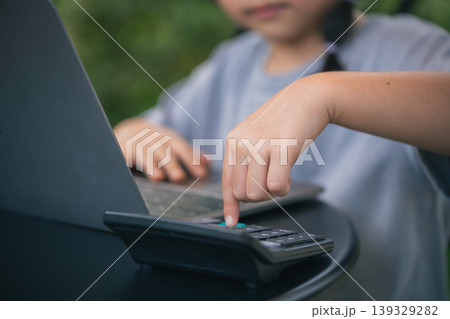 Young Child Engaged in Online Learning Using Laptop and Calculator to Enhance Skills and Knowledge in a Cozy Outdoor Setting Young Child Engaged in Online Learning Using Laptop and Calculator to Enhance Skills and Knowledge in a Cozy Outdoor Setting 139329282
