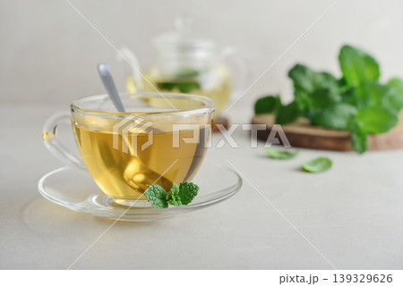 Glass cup of fresh mint tea on a light background with a glass teapot and mint leaves on a wooden board 139329626
