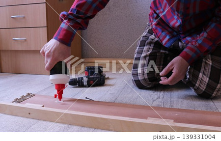 A man in a plaid suit sits on the floor in a bedroom and applies glue from a tube to the inside of a wooden bed's side plank, DIY home furniture repair 139331004