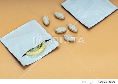 Open silver foil condom packet revealing yellow latex condom alongside scattered oval supplement tablets on warm beige background, sexual health and contraception concept photography 139331039
