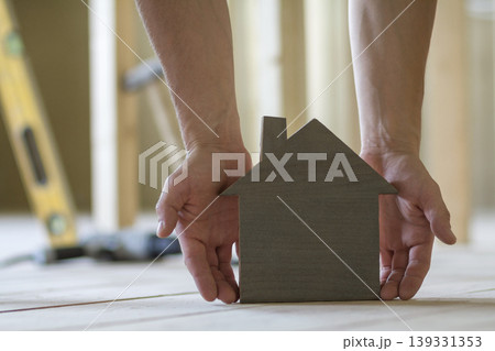 Close-up of muscular male hands holding small brown wooden model house on blurred background of building tools. Construction, investments in real estate property and ownership of dream home concept. Close-up of muscular male hands holding small brown wooden model house on blurred background of building tools. Construction, investments in real estate property and ownership of dream home concept. 139331353