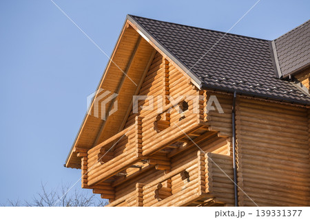 Close-up detail of new modern wooden warm ecological cottage house top with shingled brown roof and wooden sidings on blue sky background. Professionally done carpentry and construction work. Close-up detail of new modern wooden warm ecological cottage house top with shingled brown roof and wooden sidings on blue sky background. Professionally done carpentry and construction work. 139331377