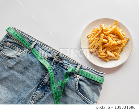 Jeans with measuring tape and fries on white background show weight loss, obesity risk and unhealthy diet concept, linking fast food temptation with body control and nutrition choices. Jeans with measuring tape and fries on white background show weight loss, obesity risk and unhealthy diet concept, linking fast food temptation with body control and nutrition choices. 139331696