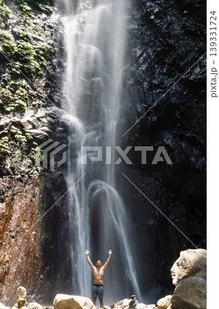 Man tourist with raised arms under powerful jungle waterfall, feeling energy in tropical nature Man tourist with raised arms under powerful jungle waterfall, feeling energy in tropical nature 139331724