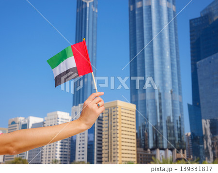 Close up of female hand holding tiny flag of UAE against Abu Dhabi skyline. Photo of UAE national day celebration spirit. Close up of female hand holding tiny flag of UAE against Abu Dhabi skyline. Photo of UAE national day celebration spirit. 139331817