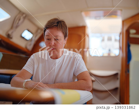 Mature woman sits inside yacht cabin analyzing paper sea chart for route planning. Navigation process involves calculating coordinates and path for upcoming sailing journey from Finike. 139331862