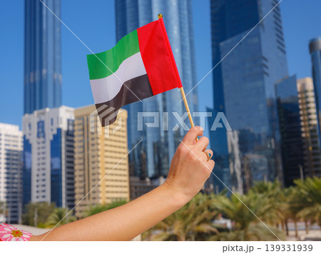 Close up of female hand holding tiny flag of UAE against Abu Dhabi skyline. Photo of UAE national day celebration spirit. Close up of female hand holding tiny flag of UAE against Abu Dhabi skyline. Photo of UAE national day celebration spirit. 139331939
