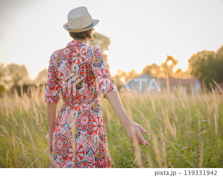 Young woman walking through picturesque European field in late summer. Golden sunlight, lush greenery, and serene rural atmosphere create peaceful countryside scene. 139331942