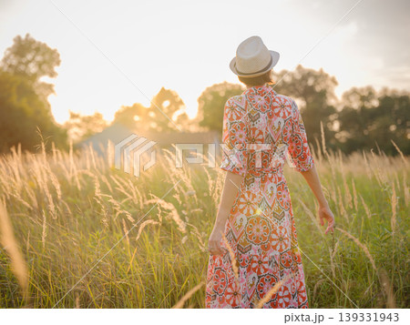 Young woman walking through picturesque European field in late summer. Golden sunlight, lush greenery, and serene rural atmosphere create peaceful countryside scene. Young woman walking through picturesque European field in late summer. Golden sunlight, lush greenery, and serene rural atmosphere create peaceful countryside scene. 139331943
