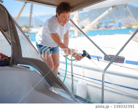 Adult woman washes teak deck of sailing yacht using water hose. Maintenance routine in Finike marina ensures boat remains clean during sunny summer day in Mediterranean Turkey. Adult woman washes teak deck of sailing yacht using water hose. Maintenance routine in Finike marina ensures boat remains clean during sunny summer day in Mediterranean Turkey. 139332391