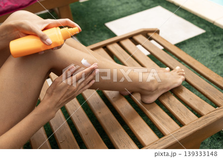 Woman applying sunscreen lotion to her leg on a wooden lounge chair outdoors by the pool. 139333148