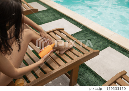 Woman applying sunscreen lotion to leg by sunny pool on a wooden lounge chair 139333150