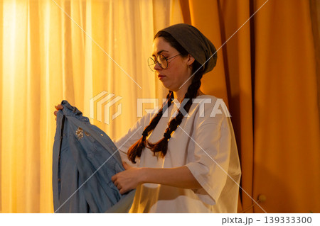 Portrait of Caucasian young woman tidying laundry in her room with evening light. She folds garments carefully. Clean interior concept. Portrait of Caucasian young woman tidying laundry in her room with evening light. She folds garments carefully. Clean interior concept. 139333300