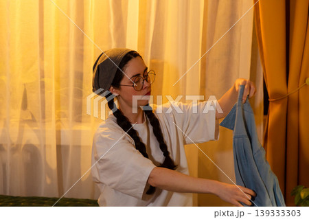 Portrait of Caucasian young woman tidying clothing in bedroom under sunset light. She keeps her room clean. Space organization concept. 139333303