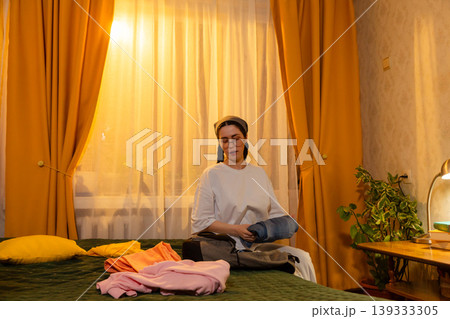 Young Caucasian woman folds clothes in her room during warm sunset light. She organizes garments indoors. Home cleaning and space organization concept. Young Caucasian woman folds clothes in her room during warm sunset light. She organizes garments indoors. Home cleaning and space organization concept. 139333305