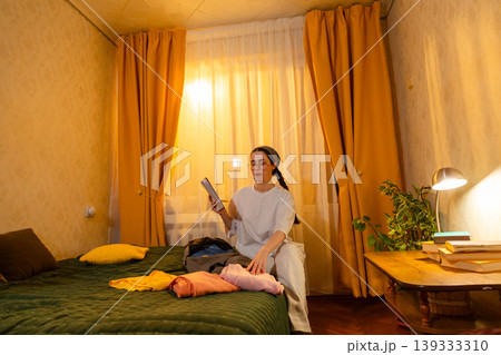 Young Caucasian woman organizing her room while folding clothes at sunset. She keeps a clean interior. Home care concept. Low angle view Young Caucasian woman organizing her room while folding clothes at sunset. She keeps a clean interior. Home care concept. Low angle view 139333310