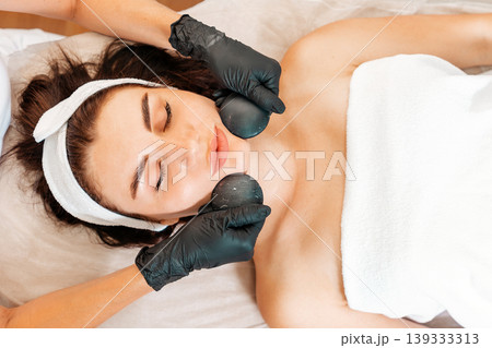 Beautician in gloves gives a facial massage with stones to the client in salon. Close up of female face. Top view. Concept of cosmetology and beauty procedures Beautician in gloves gives a facial massage with stones to the client in salon. Close up of female face. Top view. Concept of cosmetology and beauty procedures 139333313