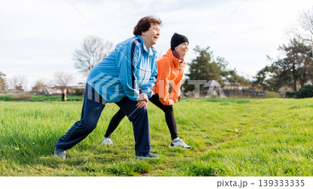 Healthy lifestyle concept. Happy grandmother and granddaughter doing sports together it the park. Old and young women in sports clothes do a stretching. Side view. International Day of Older Persons Healthy lifestyle concept. Happy grandmother and granddaughter doing sports together it the park. Old and young women in sports clothes do a stretching. Side view. International Day of Older Persons 139333335