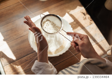 Sieving flour for cooking tasty and airy bread. Girl using sieve for flour to make it airy for baking Sieving flour for cooking tasty and airy bread. Girl using sieve for flour to make it airy for baking 139334464