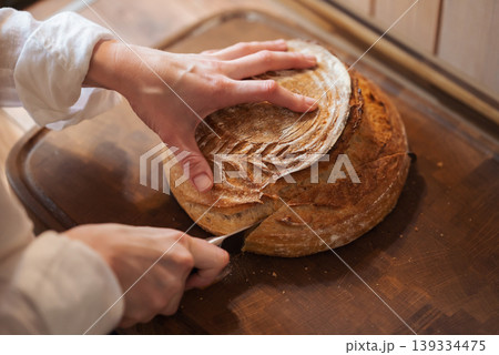 Hands of a girl cutting homemade bread on wooden tray. Baking bread at home, cutting fresh aromatic loan of bread 139334475