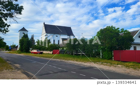 The 16th-century Catholic Church of the Holy Trinity in the Belarusian agro-town of Chernavchitsy Brest, a red car on a village road, architectural history study, rural travel spirit. 139334641