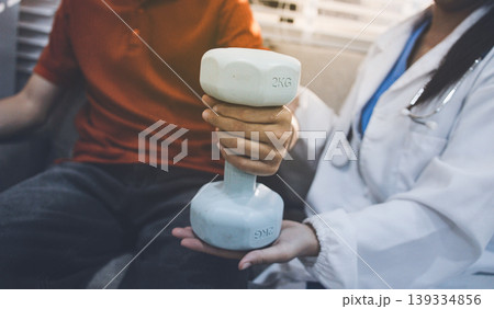 An Asian male doctor and his patient are seated in chairs, performing physical therapy using dumbbells under the doctor's supervision. The patient is experiencing pain. An Asian male doctor and his patient are seated in chairs, performing physical therapy using dumbbells under the doctor's supervision. The patient is experiencing pain. 139334856