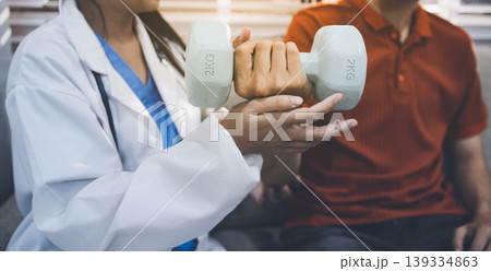 An Asian male doctor and his patient are seated in chairs, performing physical therapy using dumbbells under the doctor's supervision. The patient is experiencing pain. 139334863