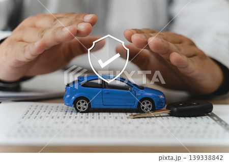 Auto insurance protection and accident prevention concept with two hands shielding blue car model, vehicle key, and policy document with shield checkmark icon, securing comprehensive coverage. 139338842