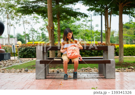 Asian student girl putting a book into her pink backpack while sitting on a park bench, Back to school and education concept. 139341326