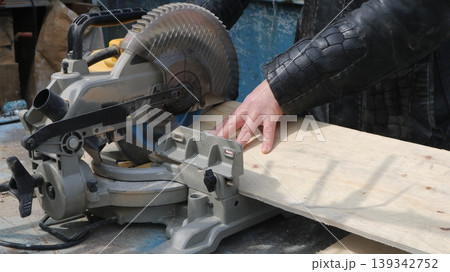 Processing a wide board on a miter saw, close-up view from the top side angle, a carpenter saws off a part of the board on the end machine 139342752
