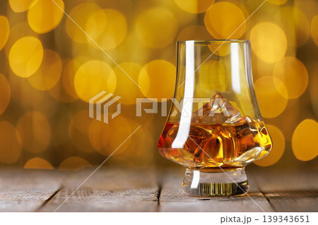 Whiskey glass containing golden amber liquor and ice cubes resting on rustic wooden surface, creating warm, elegant atmosphere with blurred bokeh background Whiskey glass containing golden amber liquor and ice cubes resting on rustic wooden surface, creating warm, elegant atmosphere with blurred bokeh background 139343651