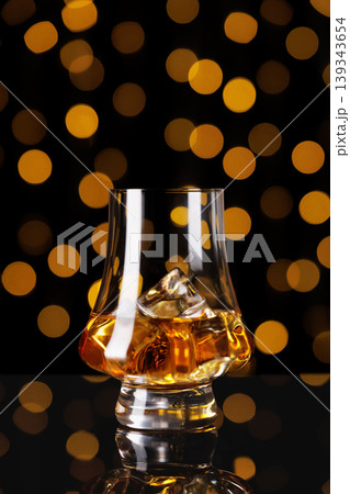 Whiskey in a tulip shaped glass with ice cubes, reflecting golden light against a dark background with orange bokeh, symbolizing a celebratory moment, tasting, or sophisticated evening 139343654