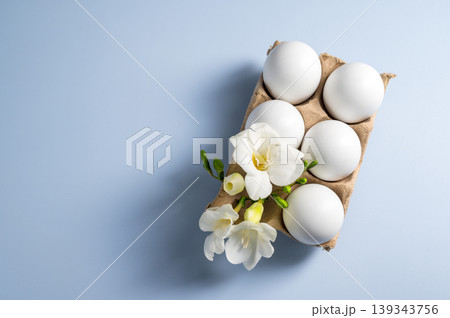 Easter eggs and flowers. White eggs in cardboard box and Freesia white spring flower, flat lay on blue background with copy space Easter eggs and flowers. White eggs in cardboard box and Freesia white spring flower, flat lay on blue background with copy space 139343756