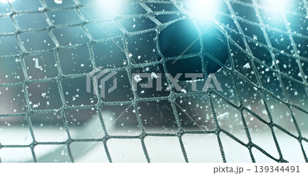 Action sports photo of a hockey puck flying into the goal net during a scoring shot, with ice particles in the air and a hockey stick visible in the background, capturing speed and intensity. 139344491