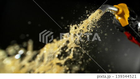 Gasoline jet spraying from fuel nozzle held by hand on dark background. Dynamic liquid motion, refueling concept, energy supply, fuel usage, dramatic lighting, macro detail. 139344521