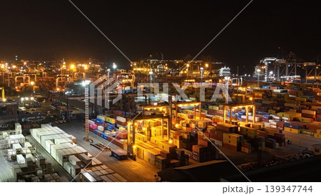 High-angle aerial view of a busy commercial container port terminal at night, featuring colorful shipping crates stacked under bright industrial lights with cranes operating in the background. High-angle aerial view of a busy commercial container port terminal at night, featuring colorful shipping crates stacked under bright industrial lights with cranes operating in the background. 139347744