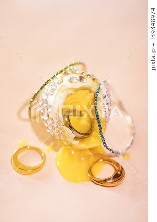Silver and gold rings alongside a broken glass bowl with yellow liquid spilling out, creating a striking contrast on a soft pink background Silver and gold rings alongside a broken glass bowl with yellow liquid spilling out, creating a striking contrast on a soft pink background 139348974