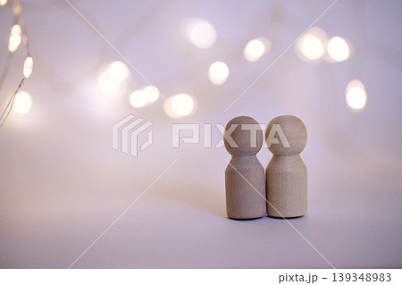 Two wooden figurines standing close together against a blurred background of warm fairy lights, creating a cozy and inviting atmosphere Two wooden figurines standing close together against a blurred background of warm fairy lights, creating a cozy and inviting atmosphere 139348983