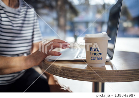 Person with striped shirt using laptop at a wooden table with coffee cup displaying text, sunlight filtering through a window in a modern cafe setting Person with striped shirt using laptop at a wooden table with coffee cup displaying text, sunlight filtering through a window in a modern cafe setting 139348987