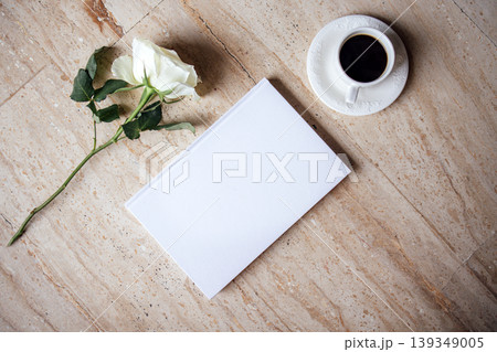 White notebook with blank pages placed beside a cup of black coffee and a single white rose on a textured stone surface, creating a serene and inviting workspace atmosphere White notebook with blank pages placed beside a cup of black coffee and a single white rose on a textured stone surface, creating a serene and inviting workspace atmosphere 139349005