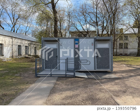 Public toilet facility with accessible entrance and WC signage stands in park area with trees and buildings. Urban infrastructure, public service, accessibility, city environment, outdoor facility Public toilet facility with accessible entrance and WC signage stands in park area with trees and buildings. Urban infrastructure, public service, accessibility, city environment, outdoor facility 139350796